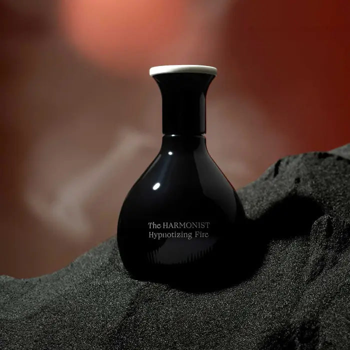 The Harmonist Hypnotizing Fire perfume bottle resting on fine black sand with a warm, smoky red background.